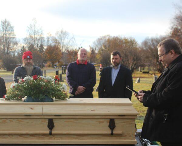 People stand around a light wooden coffin